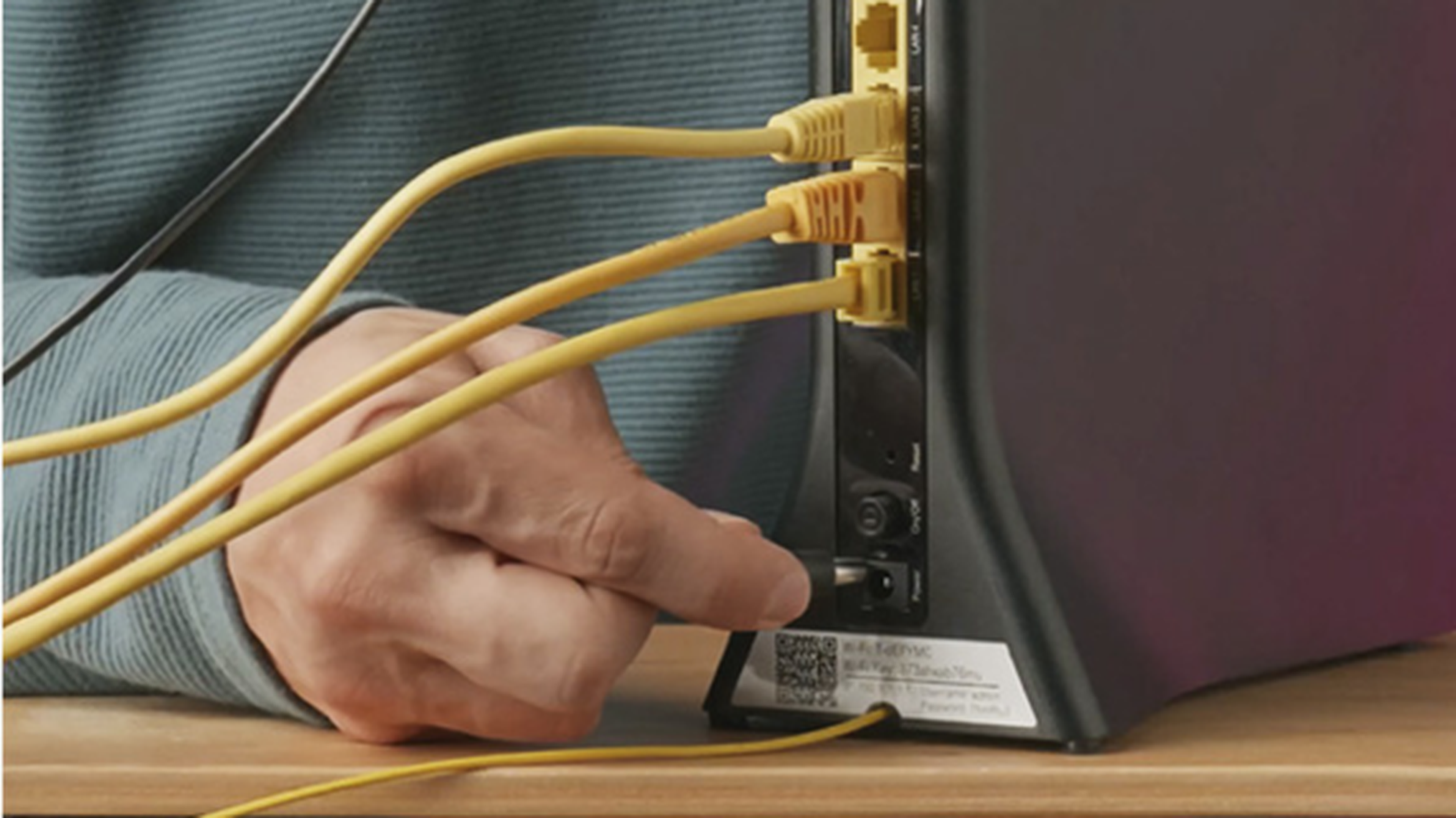 Osoba pripája žltý ethernetový kábel k čiernemu routeru na drevenom povrchu, pričom ostatné káble sú už zapojené.