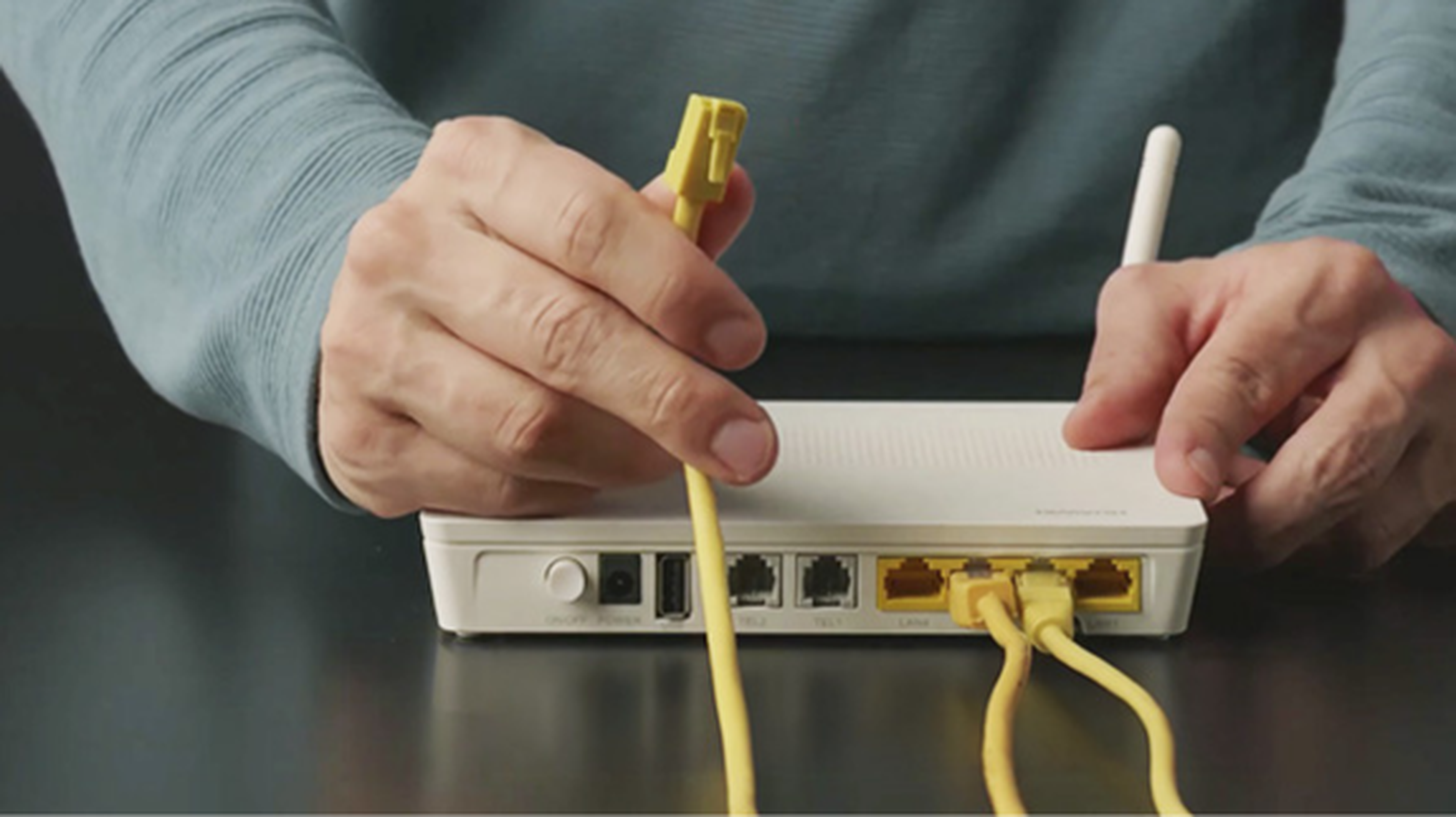 Ruky pripájajúce žltý ethernetový kábel k bielemu routeru na tmavom povrchu.