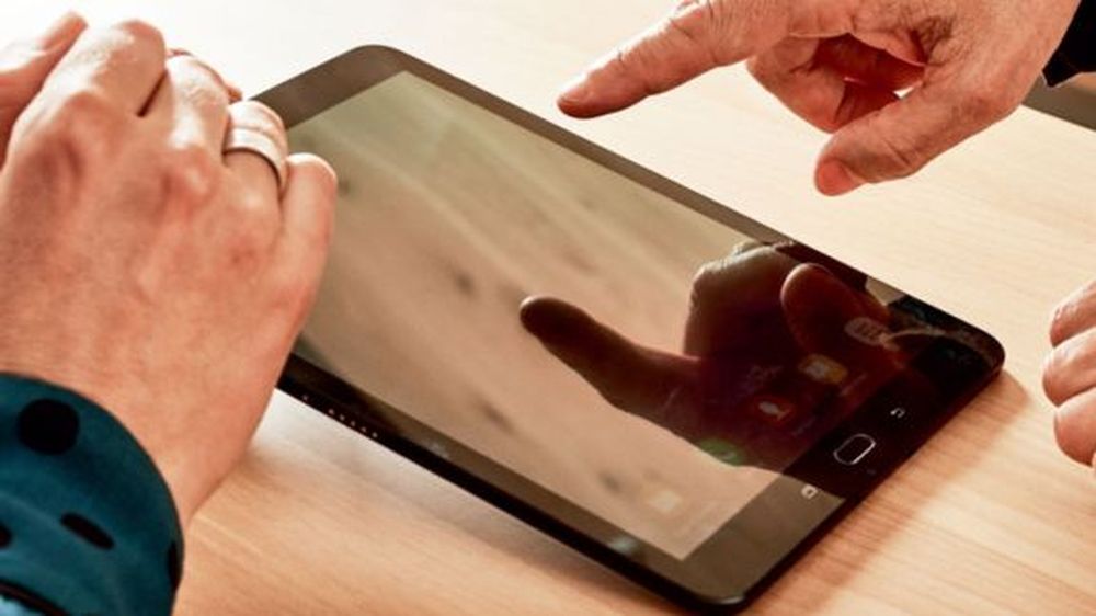 Dve ruky interagujú s tabletom na drevenom stole; jedna ruka ukazuje, zatiaľ čo druhá drží zariadenie.