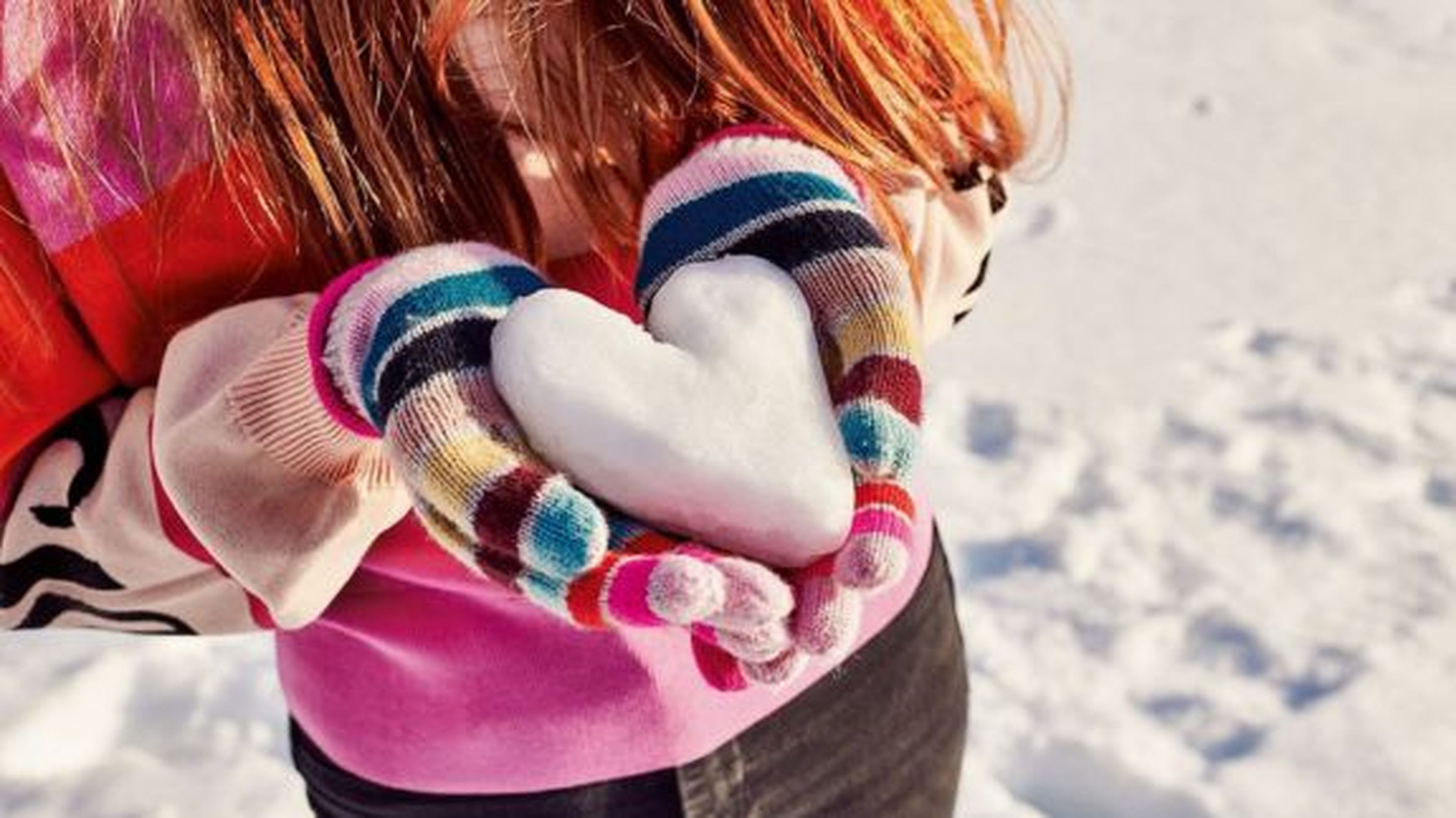 Osoba vo farebných rukaviciach drží snehovú guľu v tvare srdca a stojí na zasneženej zemi.