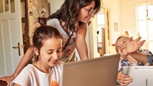 Žena sa usmieva na dve deti sediace pri stole s notebookmi, cez ktoré cez okno prúdi slnečné svetlo.
