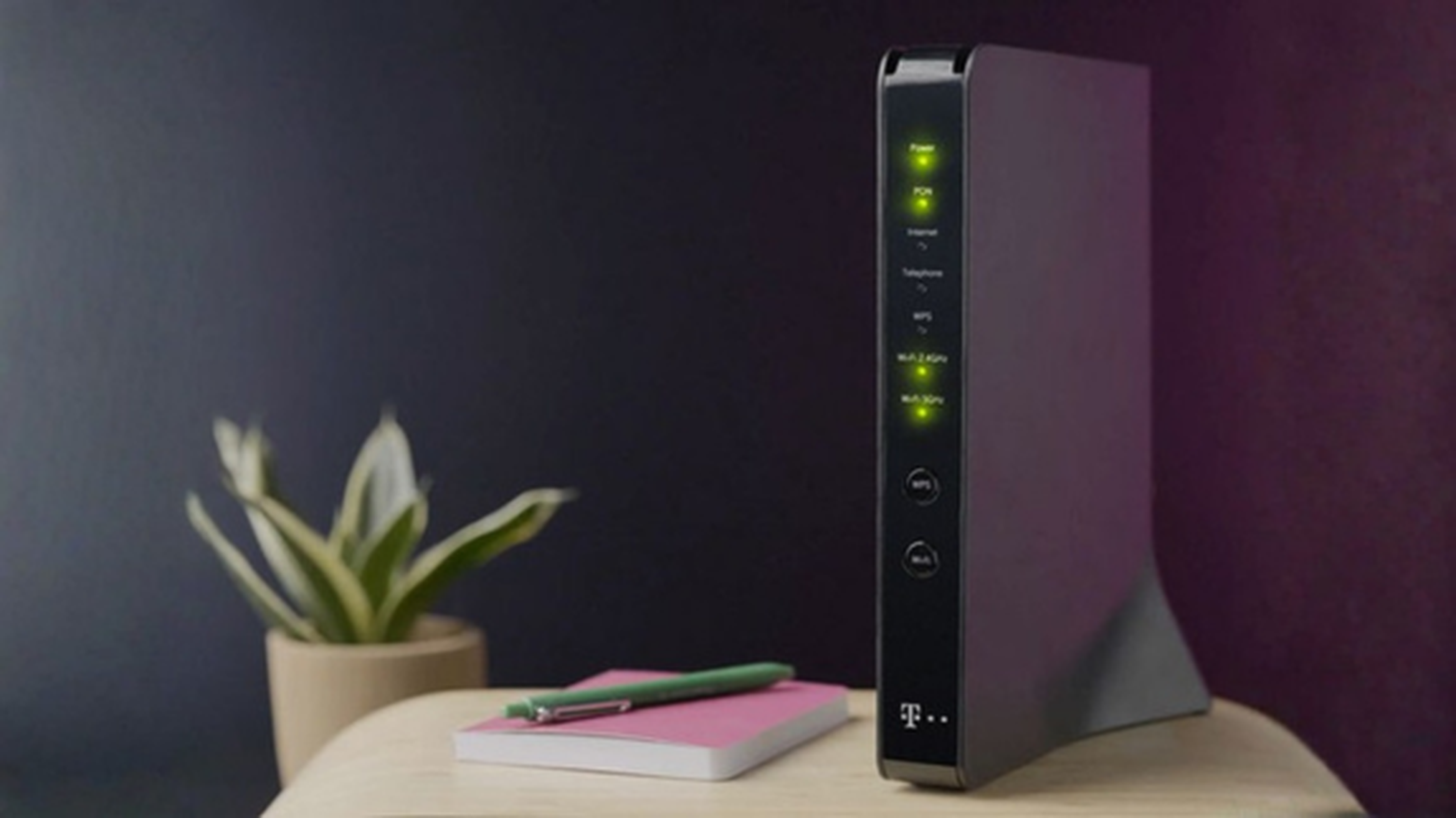 Čierny Wi-Fi router so svietiacimi kontrolkami sedí na drevenom stole vedľa malej rastliny a ružového zápisníka s perom.