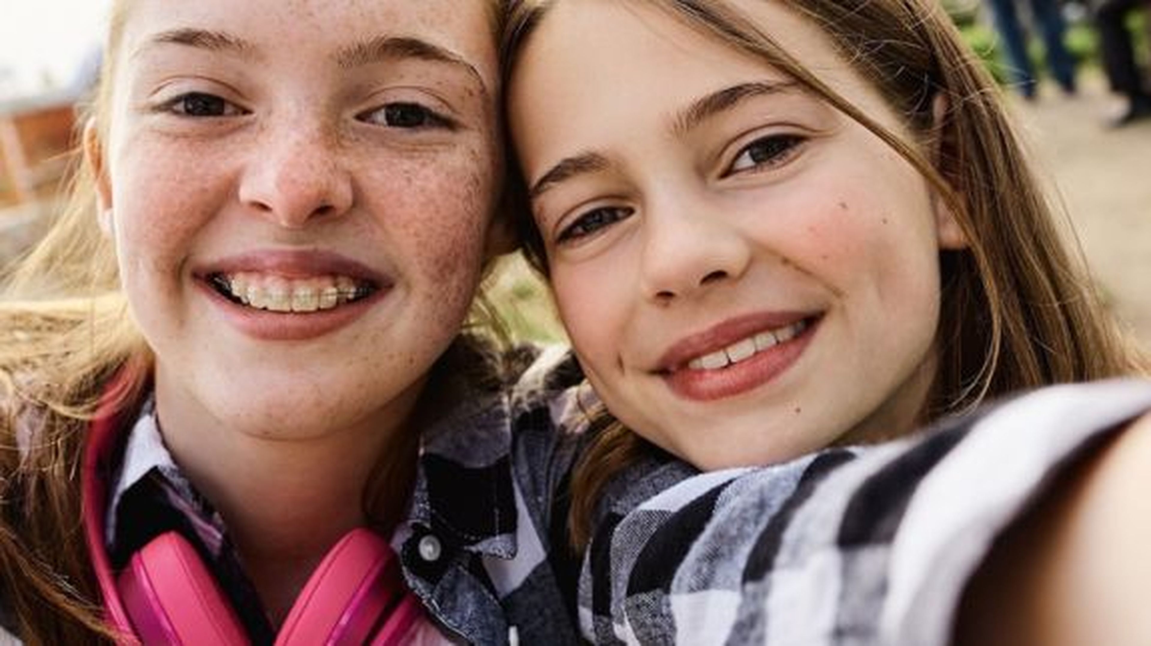 Dve usmiate dievčatá si robia selfie vonku. Jedno má pehy a strojček na zuby, nosí ružové slúchadlá a má na sebe kockovanú košeľu.