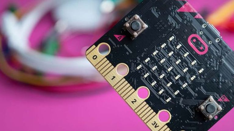 Detailný záber dosky mikrokontroléra s tlačidlami a zlatými konektormi na rozmazanom ružovom pozadí s drôtmi.