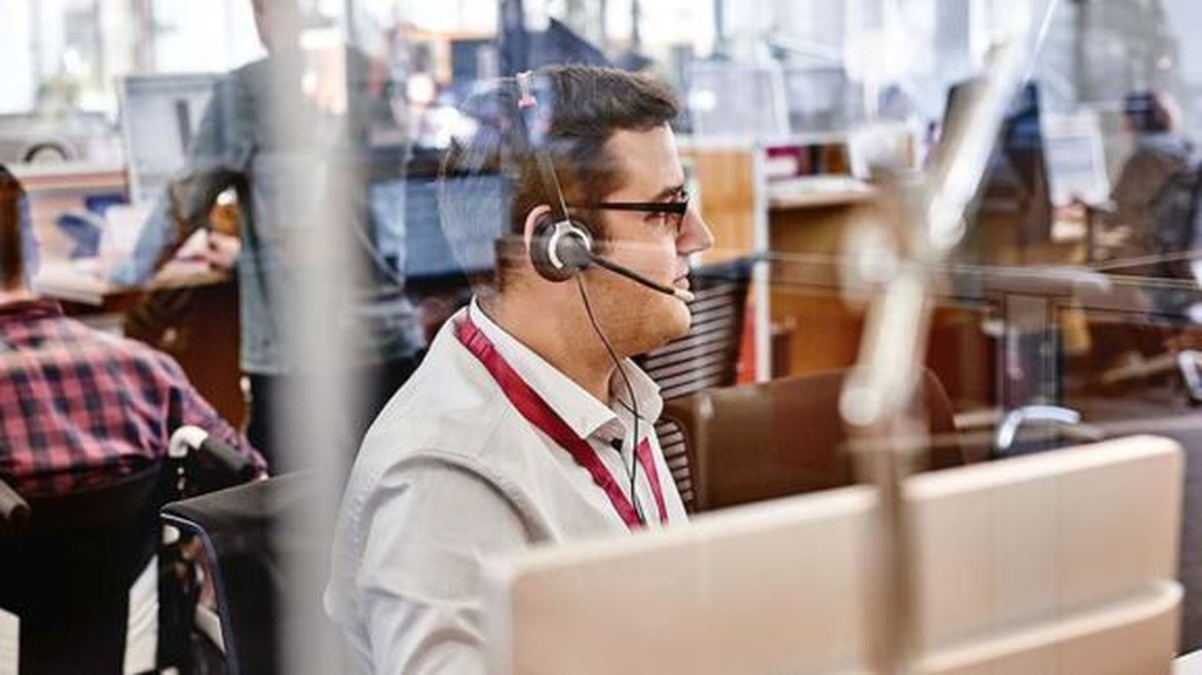 Muž s náhlavnou súpravou pracuje za počítačom v rušnom kancelárskom prostredí, videný cez sklenenú priečku.