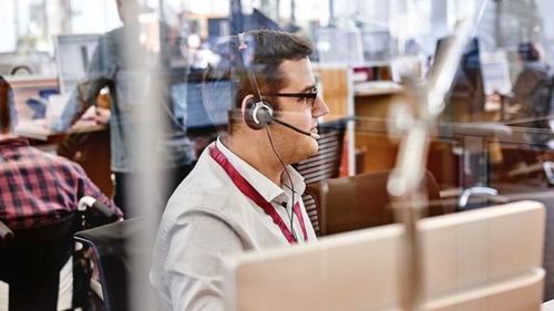 Muž s náhlavnou súpravou pracuje za počítačom v rušnom kancelárskom prostredí, videný cez sklenenú priečku.