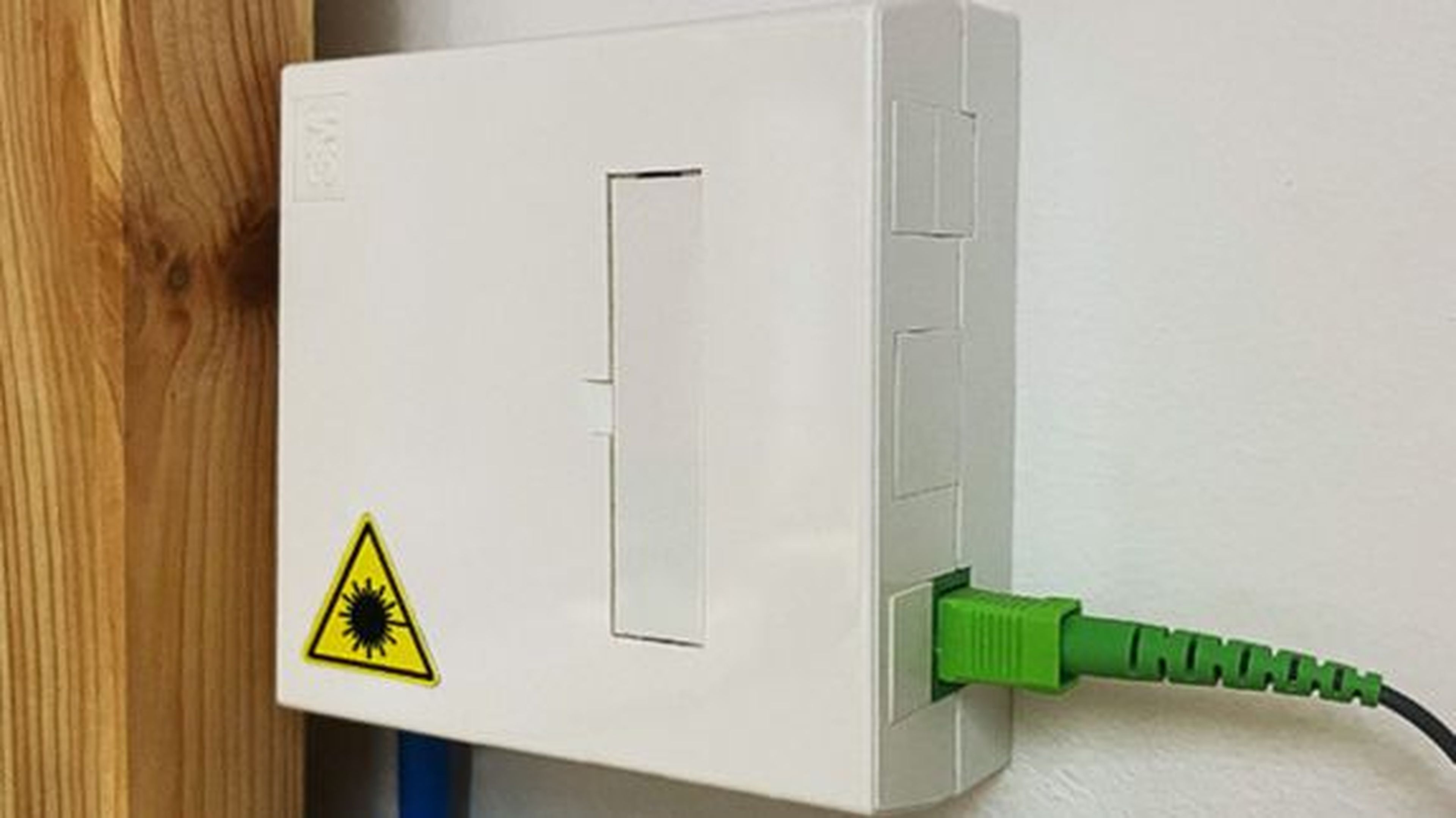 Biela nástenná optická krabica so zeleným pripojeným káblom a žltým výstražným štítkom označujúcim nebezpečenstvo laserového žiarenia.