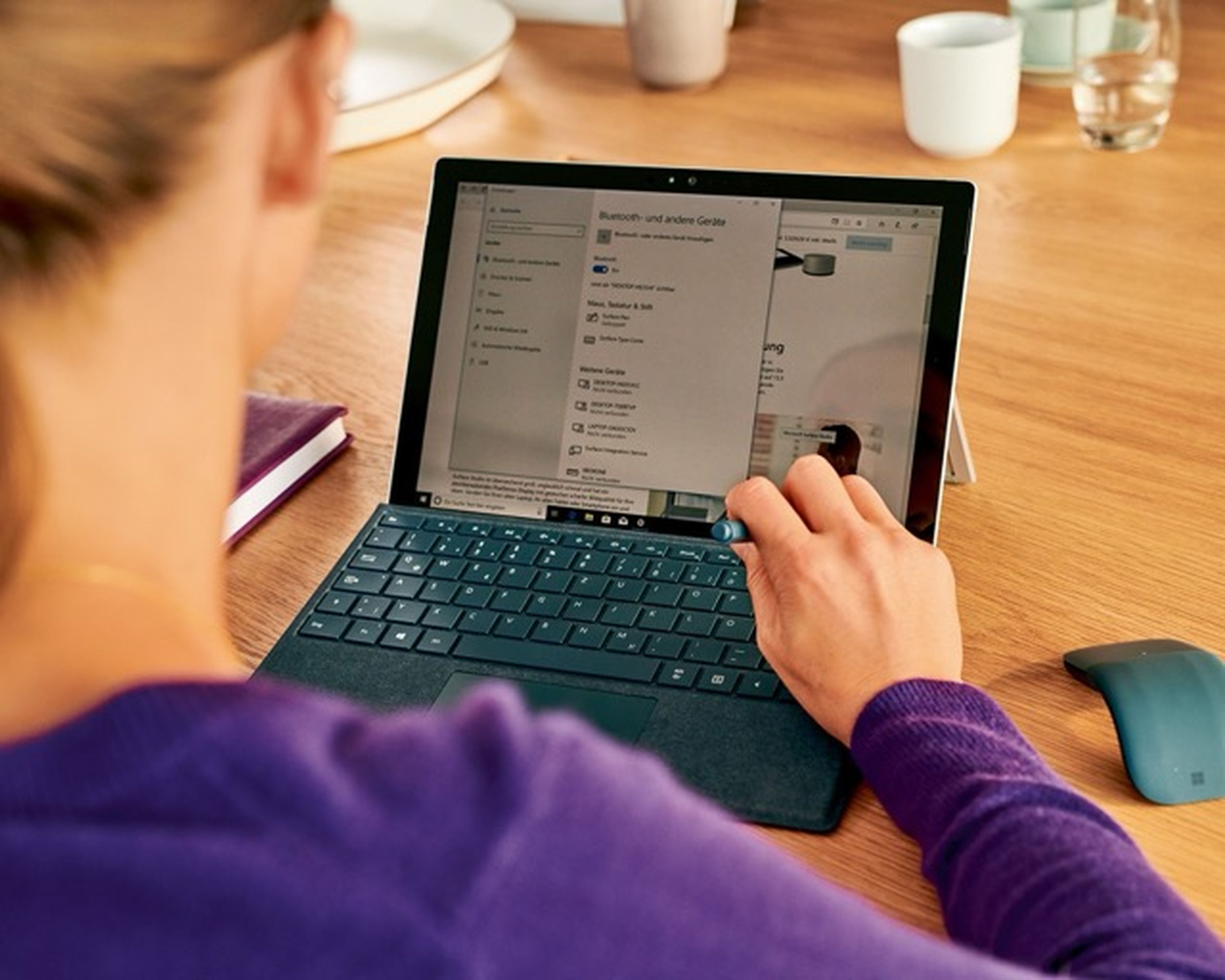 Osoba používajúca konvertibilný notebook s odnímateľnou klávesnicou na drevenom stole a prepínajúca nastavenia Bluetooth. Neďaleko je myš a dva poháre.