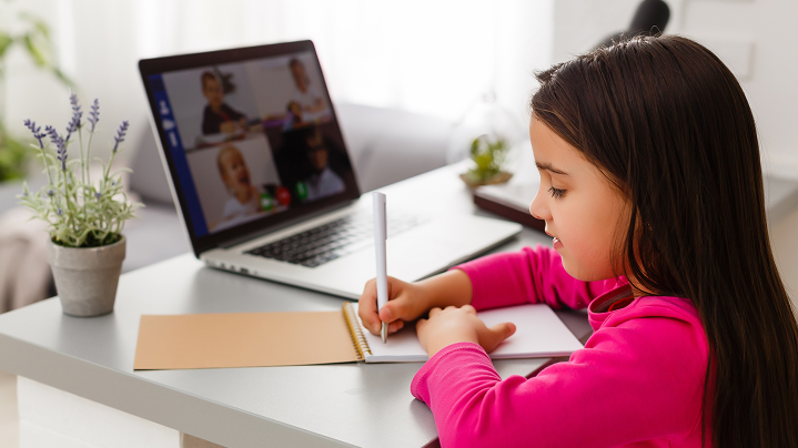 Školáčka počas online hodiny píše do zošita, na notebooku beží video hovor - zobrazuje bezproblémové online vzdelávanie cez cloud.