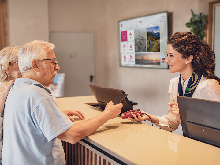 Recepčná s úsmevom podáva zákazníkovi platobný terminál - za nimi obrazovka s Telekom digitálnym obsahom. Ilustruje využitie digitálneho marketingového riešenia v praxi.