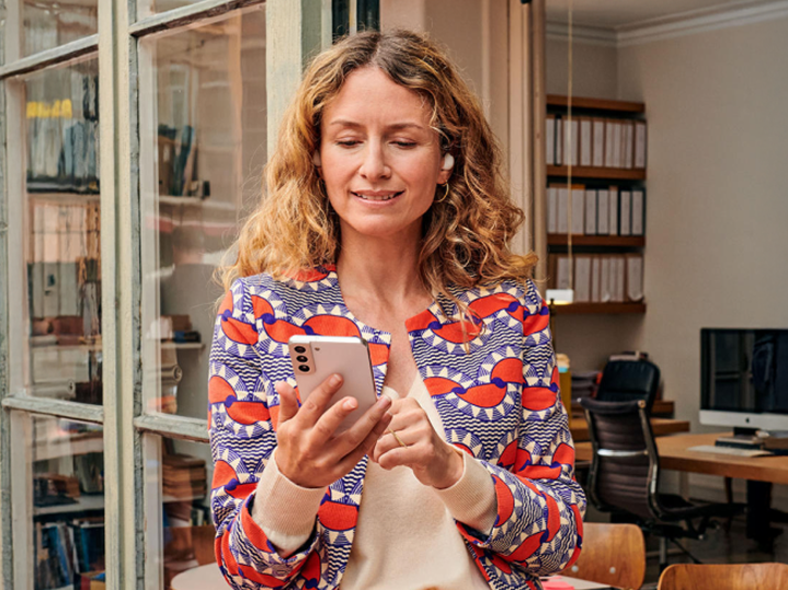 Žena v pestrofarebnom saku drží v ruke smartfón a prezerá si SMS správy. Symbolizuje efektívnu zákaznícku komunikáciu pomocou Business Messaging riešení.